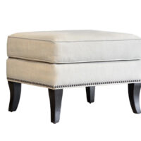 Angled shot of the Atticus Ottoman by Vogel with wooden legs, cream fabric, and nailhead trim